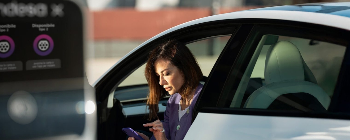 Una mujer consulta su teléfono móvil mientras permanece sentada en un coche eléctrico junto a una estación de recarga.